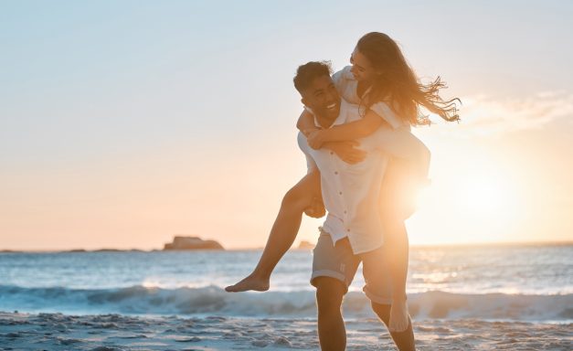 Hochzeiten Strand Ein Paar läuft am Strand bei Sonnenuntergang; der Mann trägt die Frau auf dem Rücken.